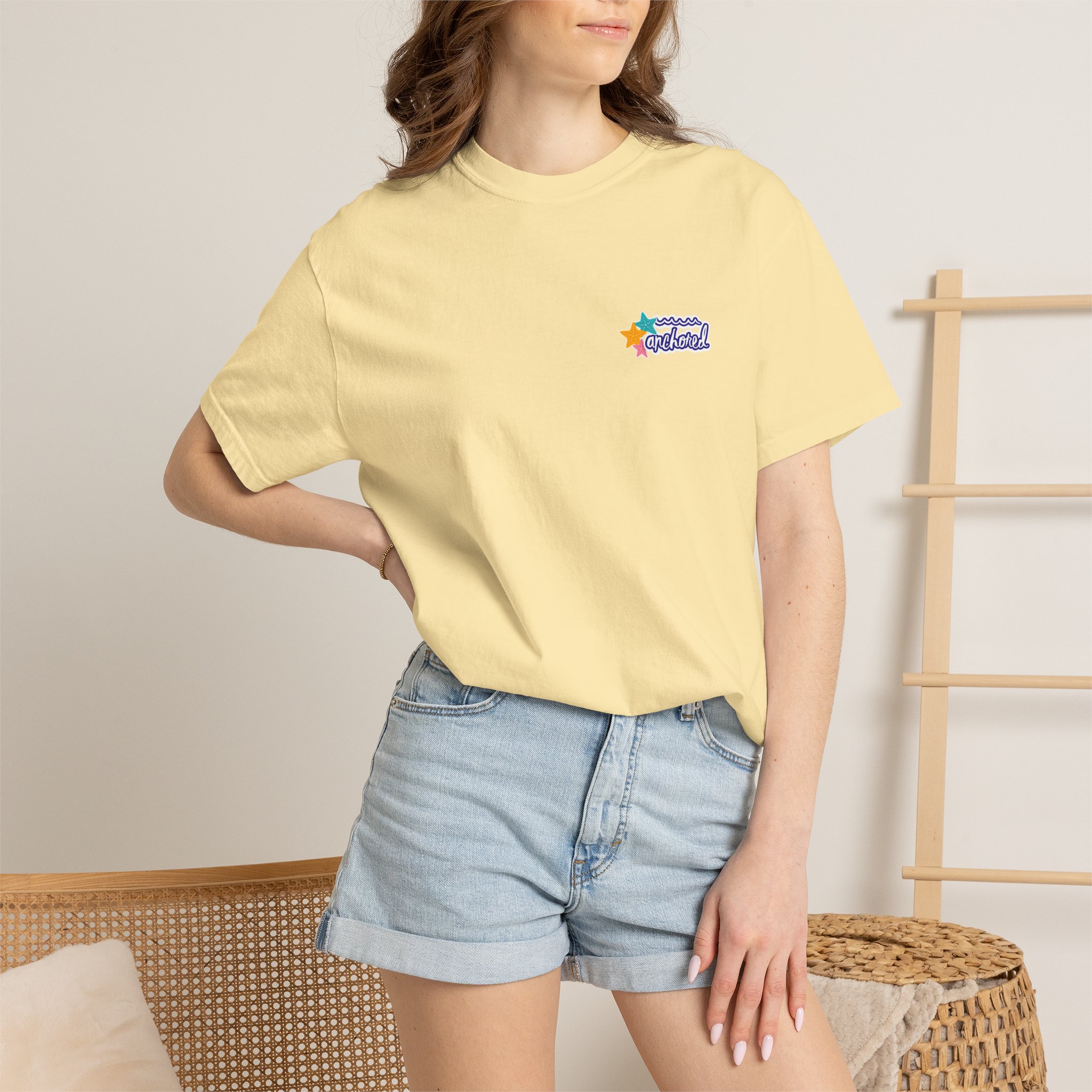 11414574105544457703_2048jpeg - Anchored Woman wearing a pale yellow T-shirt with a small colorful logo featuring stars on the left chest; she sits beside a woven basket and wooden rack, mid-torso pose.
