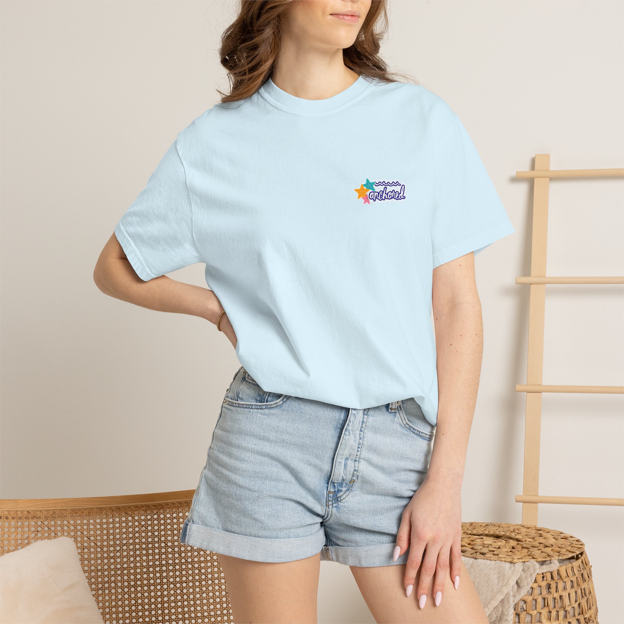 14263783579720103107_2048jpeg - Anchored Person wearing a pale blue T-shirt with a small colorful star logo on the chest, paired with denim shorts, seated beside woven furniture.