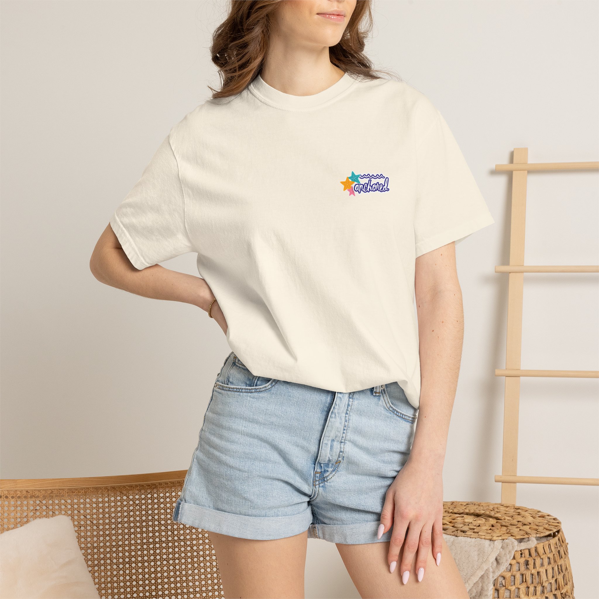 5131593909949582726_2048jpeg - Anchored Person wearing a white T‑shirt with a small colorful logo on the chest, paired with light denim shorts, sitting beside woven furniture.